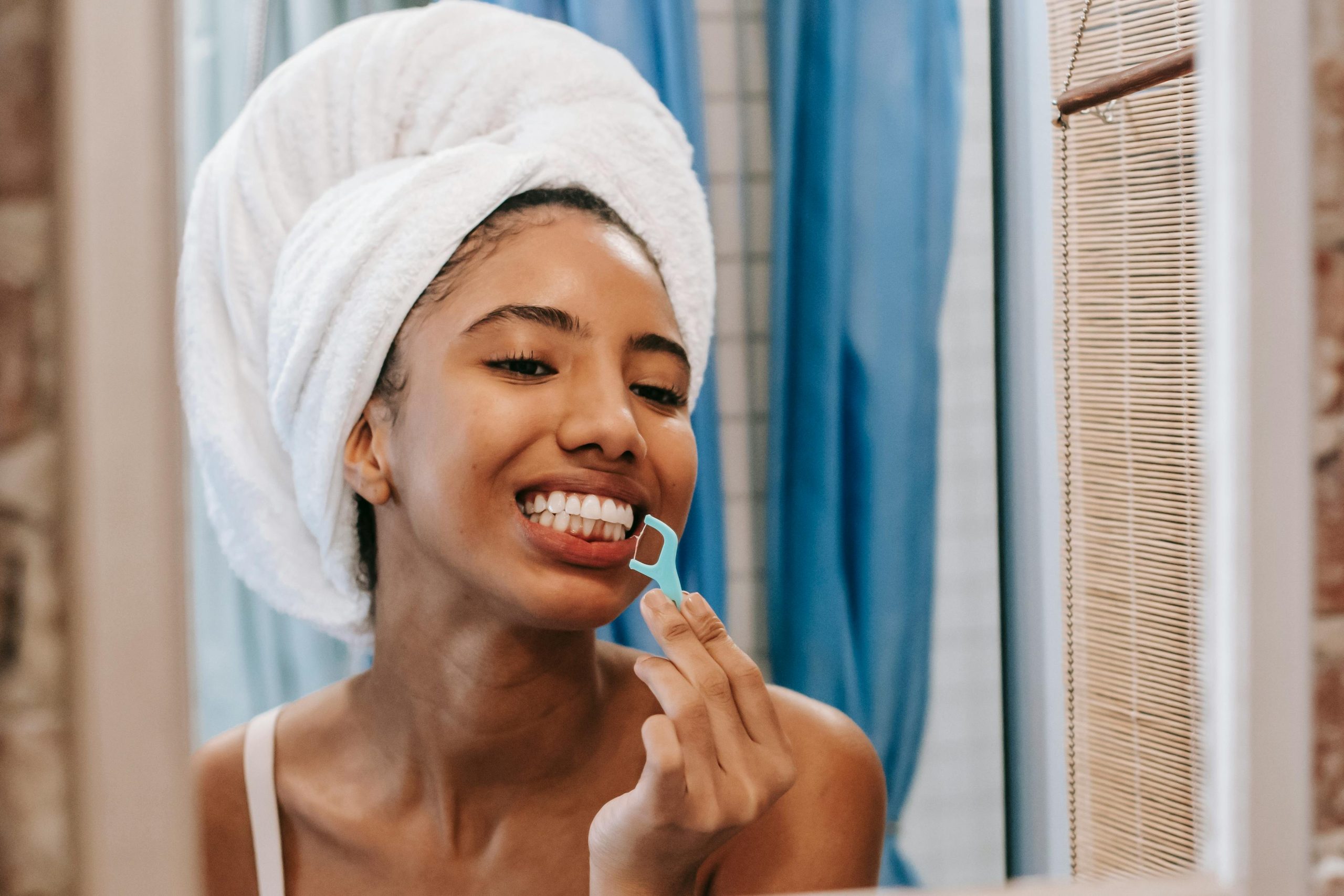 woman with a towel wrapped around her head flossing in front of a bathroom mirror